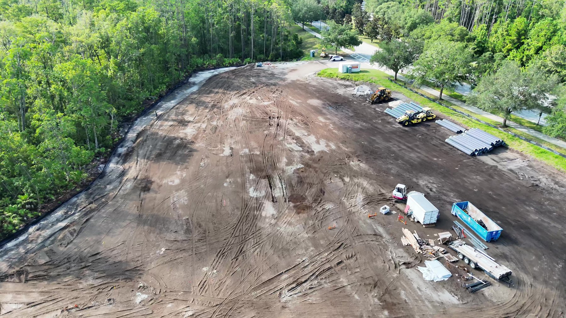 Aerial frame showing material laydown, pipe stock, and staged equipment along the eastern edge of an active construction pad.