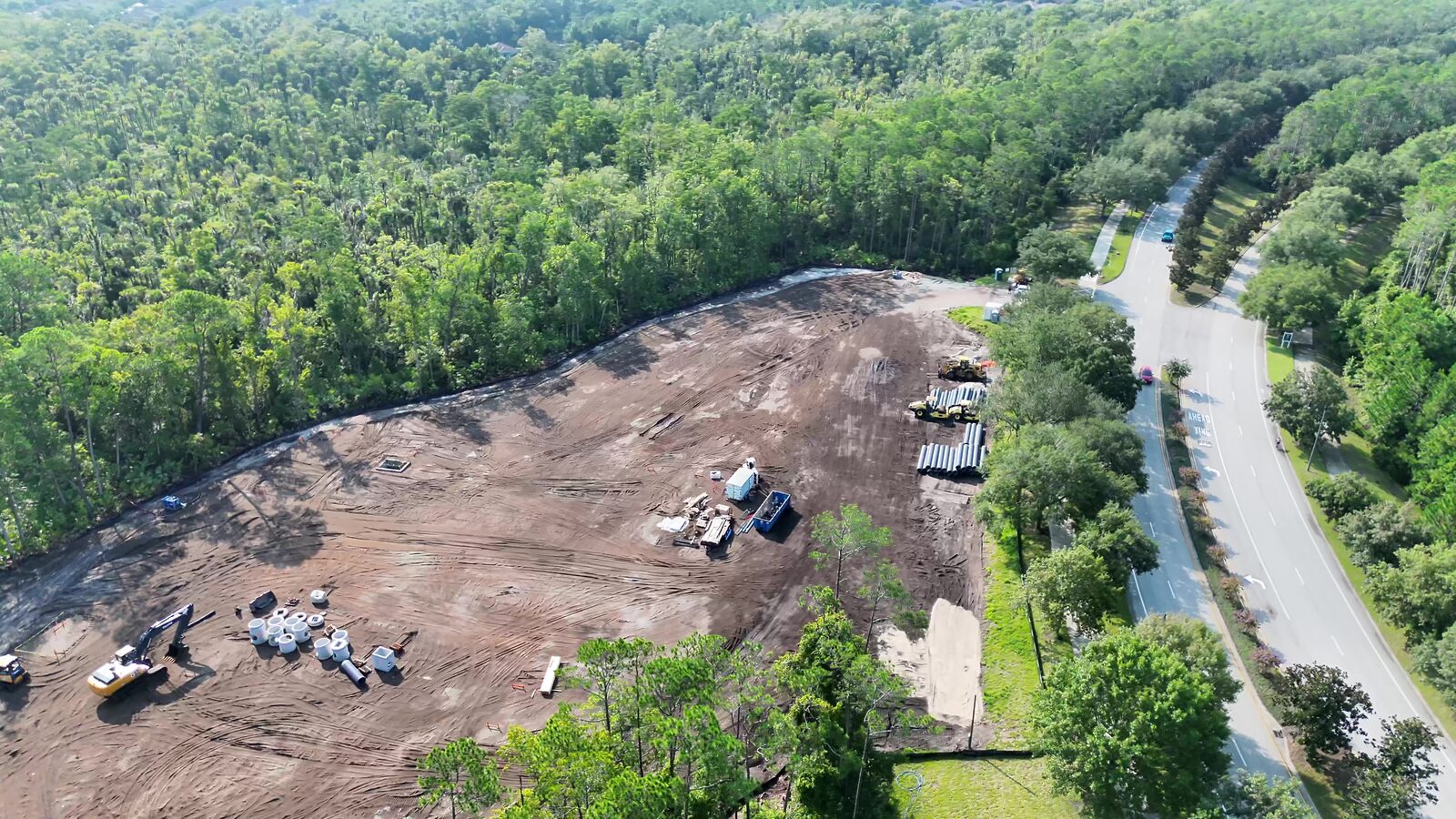 Oblique aerial view of an active Northeast Florida construction site — graded pad, staged pipe, erosion controls at the tree line, and the adjacent roadway.